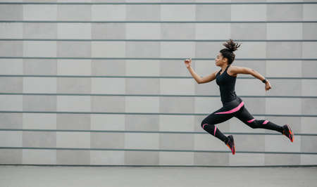 Excellent training running. African american girl in fashionable sportswear froze in air above ground, jumping on wall background, side view, panorama, free spaceの写真素材