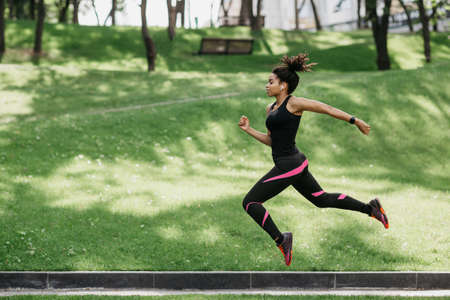 Jump and running outdoors at nature. Happy african american girl in sport uniform froze in air, in park, free spaceの写真素材