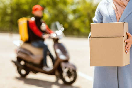 Delivery Concept. Unrecognizable Woman Holding Delivered Boxes Standing Near Courier On Scooter Outdoors. Cropped, Selective Focusの写真素材