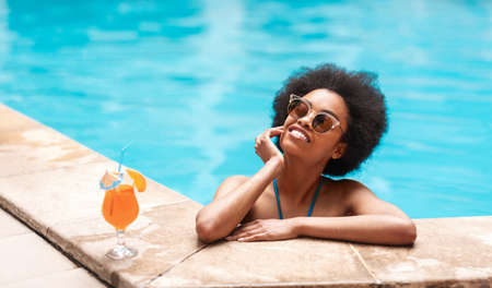 Beautiful black woman in blue bikini and sunlasses relaxing in swimming pool with cocktail drink, copy space. Panoramaの写真素材