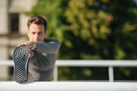 Flexibility and exercise. Muscular guy in sportswear puts his foot on railing and does stretching or exercise at park, close up, free spaceの写真素材