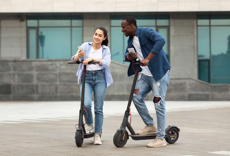 Lifestyle Concept. Cheerful diverse couple using cell phones and riding stand-up scooters, copy...