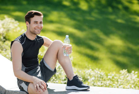Health and sports. Smiling man in sportswear holding a bottle of water and sitting in park, free spaceの写真素材