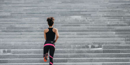 City run. Slim muscular african american girl in bright athletic uniform runs up the stairs, back, panorama, free spaceの写真素材