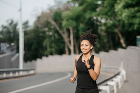 Girl runs in summer at city. Happy african american woman in sportswear with fitness tracker runs along track, free spaceの写真素材
