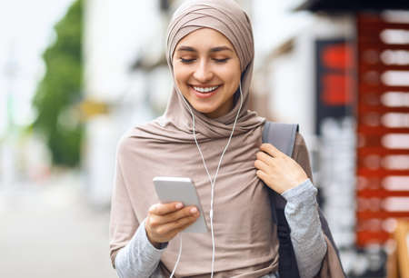 Closeup of laughing girl in hijab watching funny content on phone, walking by streetの写真素材