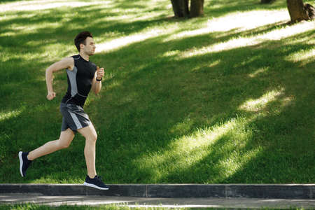 Sportsman in park. Guy in sportswear with sneakers and fitness tracker runs at track, side view,...