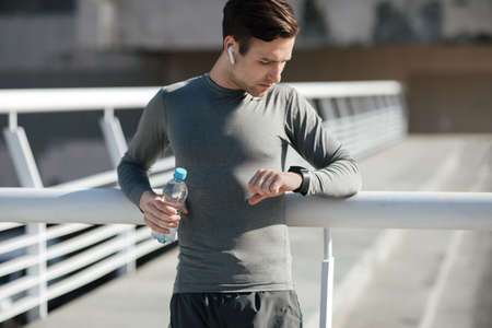 Running results. Muscular guy with headphones and bottle of water are looking at fitness tracker at...