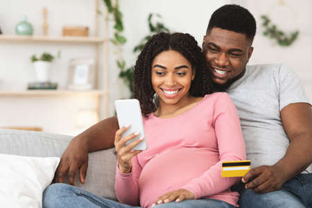 Happy pregnant black couple using mobile application for shopping online, sitting at home, using credit card, free spaceの写真素材