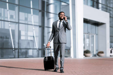 Happy African Businessman Chatting On Phone Arriving At Airport With Suitcase Standing Outside. Full Length, Free Spaceの写真素材
