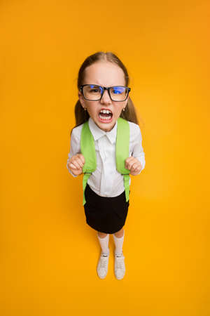 Discontented Schoolgirl In Eyeglasses Frowning Looking At Camera Standing On Yellow Studio...