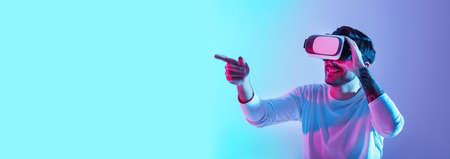 Acquaintance with virtual world. Smiling guy with glasses points finger in neon on blue and violet background, panorama, copy spaceの写真素材