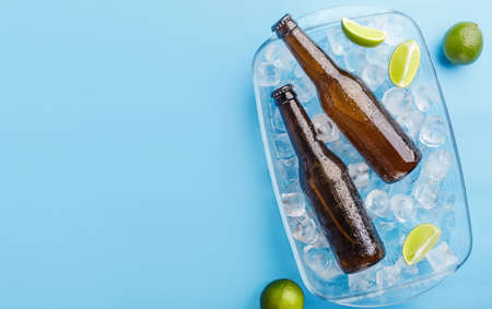 Summer, heat and beer mix. Two glass bottles without label in glass tray with ice and lime on blue background, top view, free spaceの写真素材