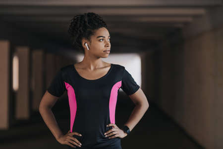 Serious african american girl in sportswear with wireless headphones and fitness tracker, outdoor,...