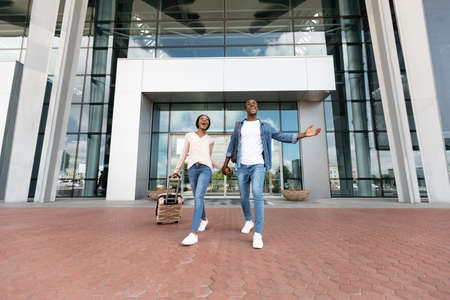 Joyful black couple leaving airport terminal with suitcases, low angle view, free space