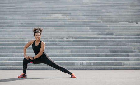 Smiling african american girl in sportswear doing stretching for legs on gray stairs background, panorama, free spaceの写真素材