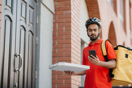 Pizza delivery with man on bike. Serious delivery man with beard in helmet, with yellow backpack holding box and looking at smartphone, near front door, free spaceの写真素材