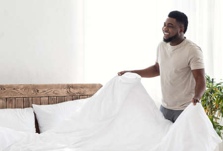 Happy black guy covering his bed with blanket and looking at empty space