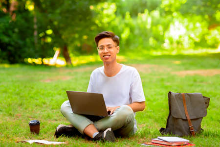 Joyful asian student guy with laptop outdoors. Smiling man sitting on lawn with computer, preparing for exams or working remotely, free spaceの写真素材