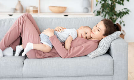 Happy mother and adorable baby boy lying on couch and embracing together, relaxing at homeの写真素材