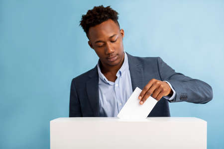 African american man putting ballot in election box over blue background, copy space