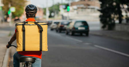 Modern fast delivery in city. Guy in safety helmet with big yellow backpack rides a bicycle on road, back, panorama, free spaceの写真素材
