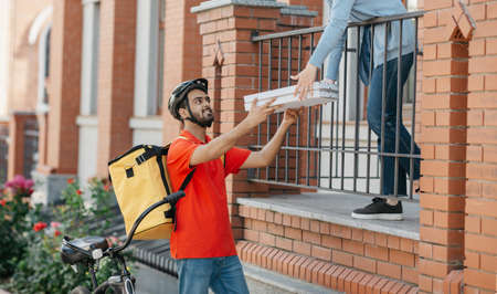 Smiling deliveryman in helmet with big yellow backpack on bicycle delivered pizza to entrance for...