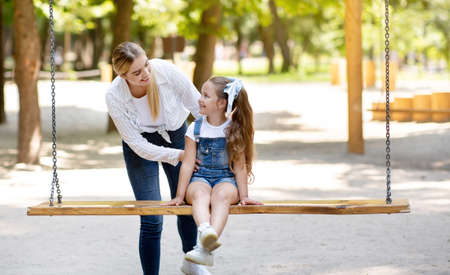 Mother Pushing Little Daughter On The Swings Spending Time Together On Playground In Park Outside....