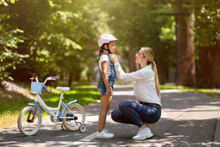Mother Putting Helmet On Daughter Teaching Her To Ride A Bicycle In Park On Weekend. Free Space For...