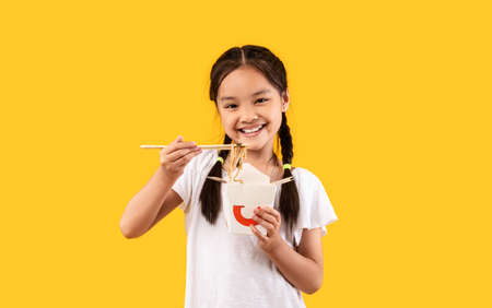 Smiling Asian Girl Eating Noodles From Box Using Chopsticks, Enjoying Chinese Food Posing On Yellow...