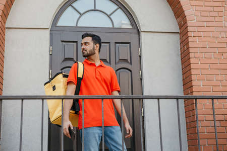 Delivering food in city courier with beard, yellow backpack and smartphone, looks for address, near...