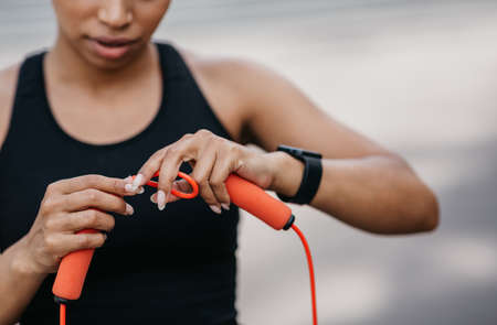 Serious african american girl in sportswear with fitness tracker adjusts skipping rope for workout,...