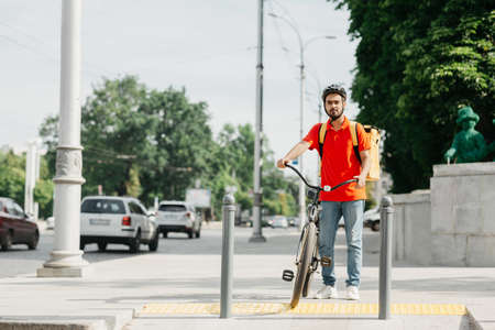 Guy with beard in protective helmet with yellow backpack and bicycle, stands at crossroads and...
