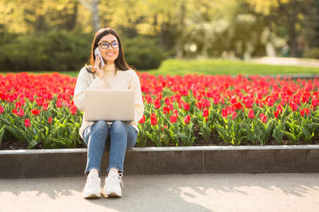 Smiling asian girl wearing glasses talking on cell phone, using laptop, sitting on parapet outdoorの写真素材