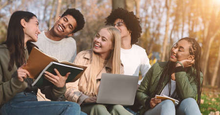 Happy teen multiracial friends doing homework together in autumn park, using laptop, taking notes,...