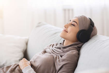 Calm Arab Girl In Hijab And Wireless Headphones Listening Favorite Songs At Home, Resting On Comfortable Sofaの写真素材