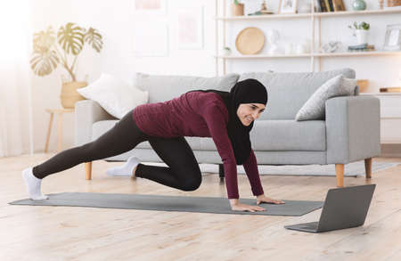 Sporty Muslim Girl Training In Front Of Laptop, Doing Leg Exercises In Living Room, Side View With Copy Spaceの写真素材