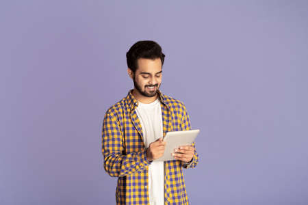 Smiling Indian man studying or working online on tablet computer against lilac background