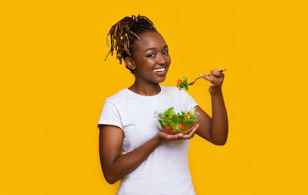 Healthy african american young woman eating fresh salad over yellow studio background, healthy food...
