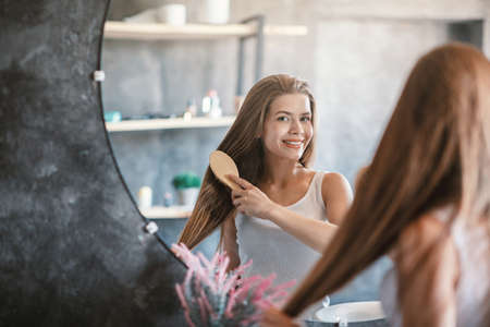 Young woman brushing her beautiful long hair with wooden brush near mirror indoors, free spaceの写真素材