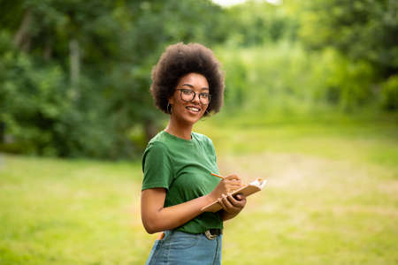 Portrait Of Cheerful Black Student Girl With Book In Hands Posing Outdoors, Taking Notes, Preparing...