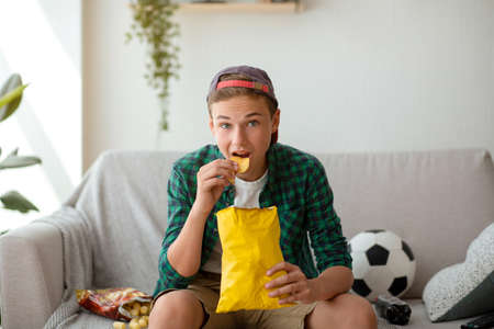 Concentrated teen guy eating chips and watching football game on TV at home