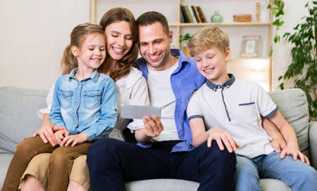 Family Of Four Using Mobile Phone Watching Cartoons Together Sitting On Sofa At Home. Weekend...