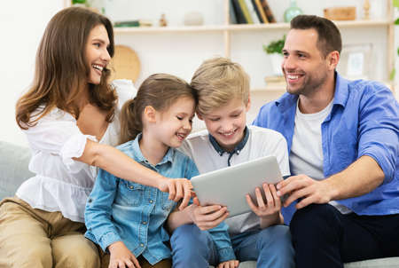 Parents And Children Using Tablet Computer Sitting On Sofa Watching Film Together At Home. Family...