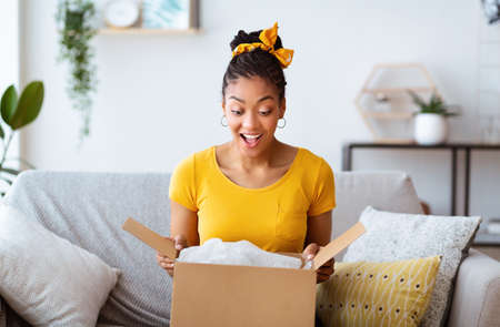 Buying Via Internet. Excited afro girl sitting on sofa unboxing cardboard delivery package, copy...