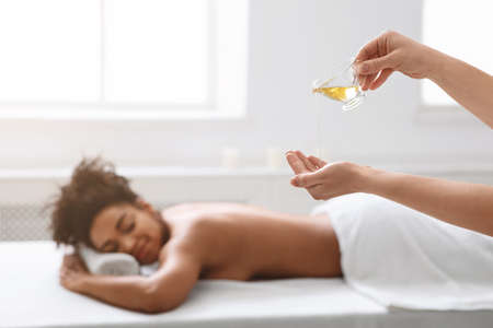 Massage therapist pouring oil on her hands, sleeping black lady waiting for massage, spa interiorの写真素材
