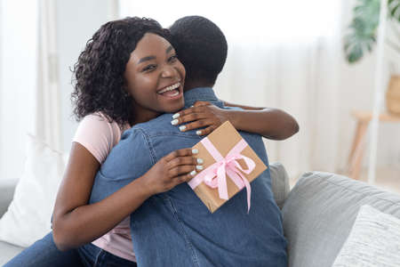 Happy black girl with gift box in her hands hugging her husband, home interior, empty spaceの写真素材