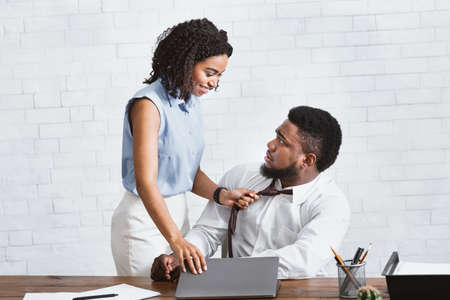 Sexual molesting at work. Black woman pulling her subordinate by his tie in company office. Panorama