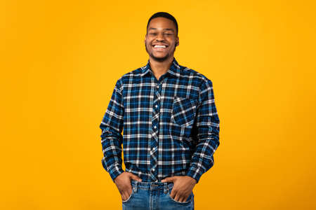 Black Millennial Guy Smiling To Camera Standing Holding Hands In Pockets Posing Over Yellow Background. Studio Shotの写真素材