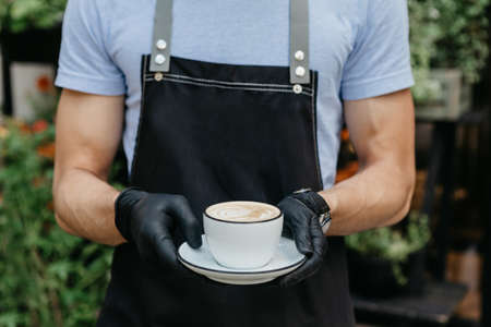 Morning coffee cup. Waiter in apron and black gloves, holding cappuccino at cafe outdoor, cropped, close up, free spaceの写真素材